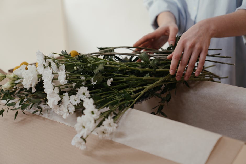 A person’s hands gently arrange a bouquet of fresh white and yellow flowers, including carnations and daisies, on a light-colored table. The bouquet features vibrant yellow blooms contrasting with t