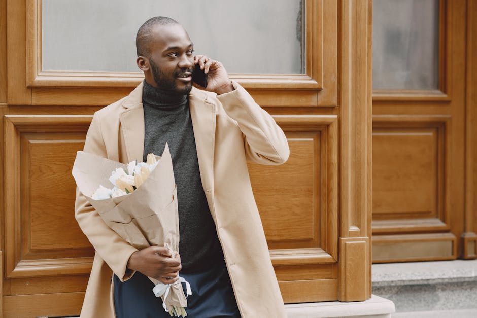 A man with a dark beard and short hair, dressed in a beige coat over a dark sweater, is standing outside in front of wooden paneling, holding a bouquet of fresh white flowers wrapped in white paper. H