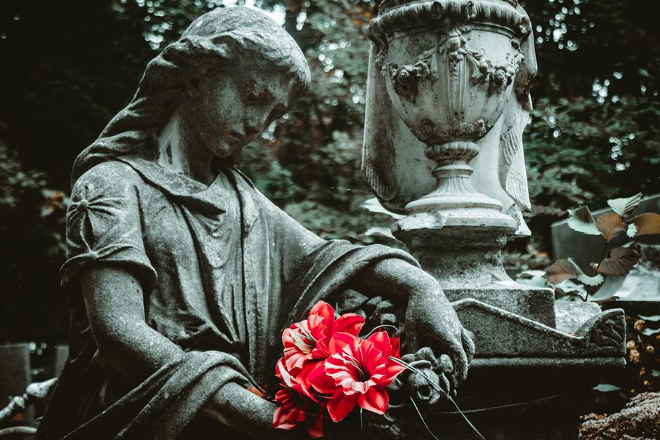 A weathered stone statue of a grieving woman holding a bouquet of vibrant red and white flowers, situated in a cemetery with surrounding urns and lush greenery. The statue's textured surface and detai
