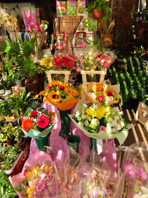 A vibrant display of fresh floral bouquets arranged on a pink cloth-covered table inside a flower shop, featuring a variety of bright-colored flowers including yellow sunflowers, pink gerbera daisies,