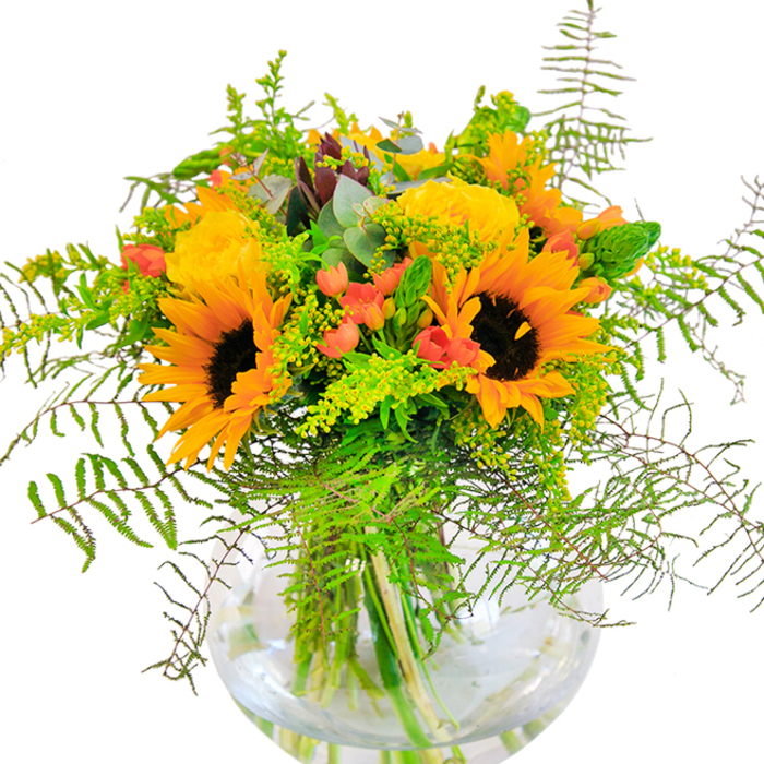 From the first glance, this bouquet exudes exuberance and sunlit optimism, embodying the lively character of Southfields' neighbourhoods. Prominently, large sunflowers face outward, their petals radiating golden warmth around deep mocha centres-a vivid nod to late August afternoons spent in Coronation Gardens. Nestled amongst them, classic yellow garden roses blossom in closely packed spirals, their velvety softness and sunny hue bridging tradition with contemporary style. Glossy red Hypericum berries punctuate the arrangement, sparkling like rubies among the green and imparting a sense of playfulness. Feathery ferns and airy sprigs of solidago infuse texture and movement, reaching outward as if touched by a gentle Southfields breeze, while hints of eucalyptus add a subtle contrast both in colour and fragrance. The stems are displayed in a clear, gently rounded vase-crystal transparency enhancing each vibrant element and recalling the freshness found in local flower stalls by Replingham Road. Crafted with the expertise of a Southfields florist, this bouquet offers a perfect gesture for celebrating birthdays, anniversaries, or simply bringing a joyous edge to everyday living within SW18 and beyond.
